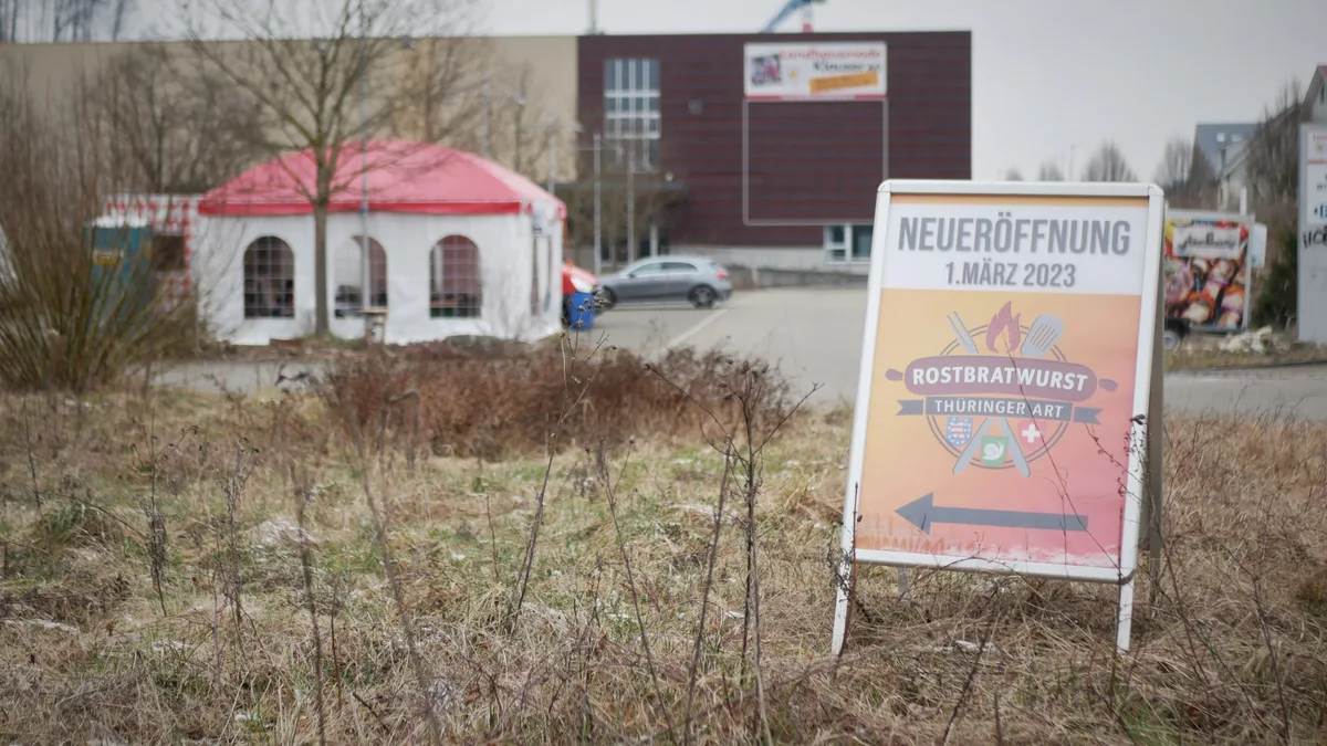 Man sieht eine orange-rote Hinweistafel auf einem Rasenstreifen, dahinter ein Partyzelt mit rotem Dach.